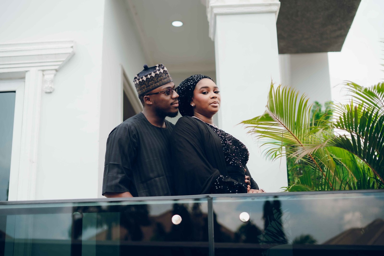 Couple in black traditional Nigerian attire standing on balcony with lush plants.