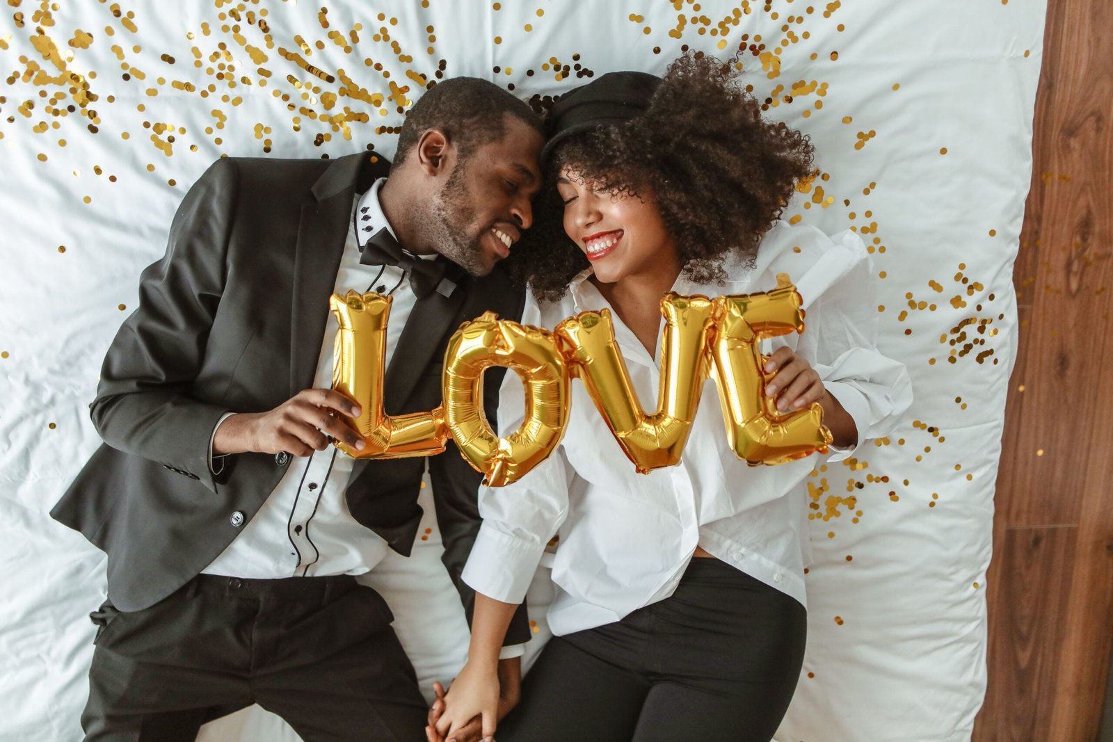 Smiling black couple lying on bed with LOVE balloons, joyful and intimate moment.